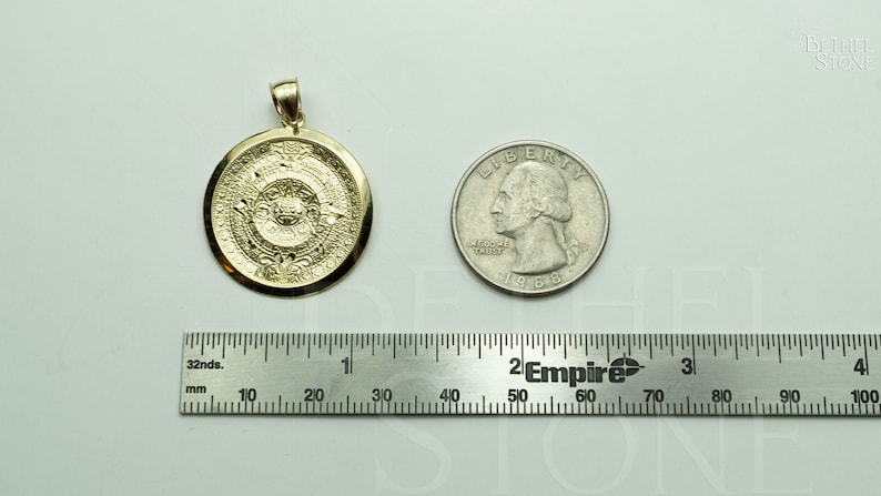 May include: A gold pendant featuring a detailed depiction of the Aztec calendar, the Sun Stone, with intricate carvings. The pendant is shown next to a US quarter for size comparison.