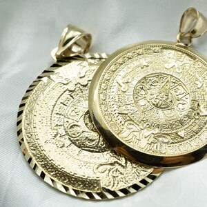 May include: Two gold pendants with intricate designs. The pendants feature a circular shape with a raised, detailed pattern. The pendants are connected by a loop at the top.