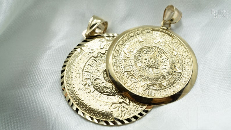 May include: Two gold pendants with intricate designs. The pendants feature a circular shape with a raised, detailed pattern. The pendants are connected by a loop at the top.