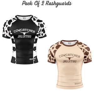 Puede incluir: Dos rashguards de manga corta. Uno es negro con manchas de vaca blancas en las mangas y los hombros, y el texto "COWCATCHER JIUJITSU". El otro es beige con manchas de vaca marrones y el mismo texto.