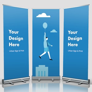 May include: Three blue retractable banner stands with silver bases. The center banner features a cartoon illustration of a person holding a balloon and floating above a building. The text "Your Design Here" appears on each banner with the tagline "Urban Sign & Print".