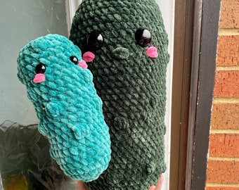 May include: Two crocheted plush toys shaped like green and blue cucumbers. The green cucumber has a pink mouth and black eyes. The blue cucumber has a pink mouth and black eyes.