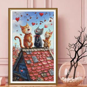 Könnte beinhalten: Gerahmtes Kunstwerk mit drei singenden Katzen auf einem roten Ziegeldach unter blauem Himmel mit Noten und Herzen. Die Katzen sind orange, grau und gelb, mit einem detaillierten, farbenfrohen Illustrationsstil. Das Kunstwerk steht auf einem Holztisch.