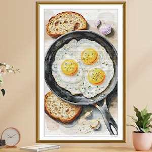 Puede incluir: Una obra de arte enmarcada representa una escena de desayuno. Tres huevos fritos en una sartén negra están rodeados de dos rebanadas de pan tostado y dientes de ajo. La obra de arte está en un marco dorado.