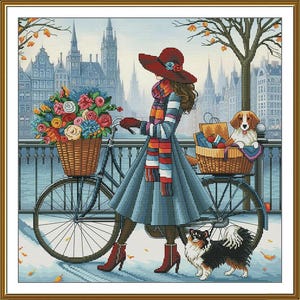 Op de afbeelding: Een kruissteekontwerp toont een vrouw met een rode hoed en sjaal die een fiets met bloemenmanden duwt. Een hond zit in de achterste mand en een andere hond staat naast de fiets. De achtergrond toont een stadsgezicht met gebouwen en bomen.