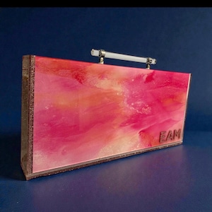 May include: A rectangular clutch with a vibrant pink and orange abstract design. The clutch has a clear handle and the letters "FAM" are visible on the bottom right corner. The edges are outlined with a glittery brown material.