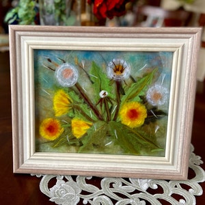 May include: A framed needle felted artwork depicting a cluster of dandelion flowers in various stages of bloom. The flowers are rendered in shades of yellow and white, with some dandelion seeds floating in the air. The background is a soft blue and green.