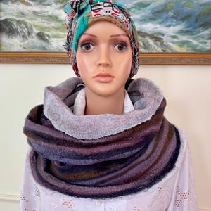 Könnte beinhalten: Mehrfarbiger Rundschal in Lila-, Braun- und Grautönen. Der Schal ist aus einem weichen, strukturierten Material gefertigt und wird um den Hals getragen. Der Schal wird auf einer Schaufensterpuppe mit gemustertem Kopftuch und weißem Hemd präsentiert.