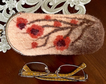 May include: A pink and brown felt eyeglass case with a floral design. The case has a metal clasp and is decorated with a brown branch with red flowers.