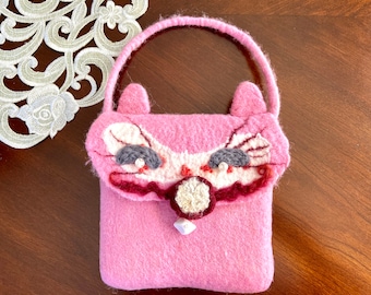 May include: A pink felt purse with a cat face design. The cat has embroidered eyes, a nose, and whiskers.