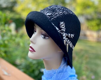 May include: A black felt cloche hat with a patterned design and a black and white ribbon accent. The hat has a wide brim and a slightly rounded crown.