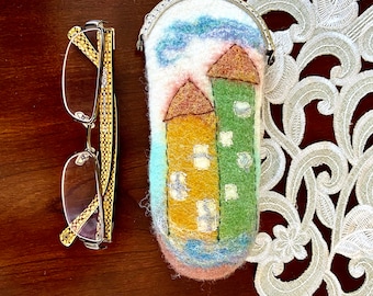 May include: A felt eyeglass case with a colorful house design. The case has a metal clasp and is decorated with a blue sky, white clouds, and green grass.