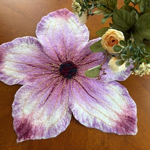 Könnte beinhalten: Ein filzförmiger Untersetzer in Form einer Blume in Lila- und Weißtönen mit goldenen Fadenakzenten. Die Mitte ist ein tiefes Burgunderrot, und die Blütenblätter haben eine strukturierte Optik. Im Hintergrund ein Blumenarrangement mit cremefarbenen und pfirsichfarbenen Rosen.