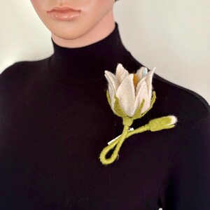 May include: A white felt flower brooch with a yellow center and green stem and leaves. The brooch is attached to a black turtleneck. The flower has a realistic design, with detailed petals and a delicate appearance.