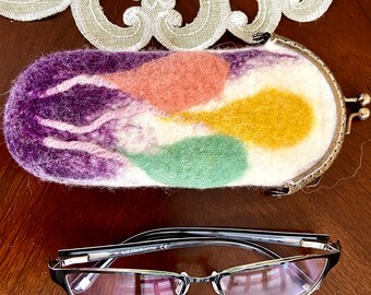 May include: A white felt eyeglass case with a colorful abstract design in shades of purple, pink, green, and yellow. The case has a metal clasp and is sitting on a wooden surface. A pair of black framed eyeglasses are also pictured.