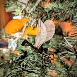 May include: A bronze-colored key ornament with delicate, translucent wings, suspended by a gold chain. The key is detailed with ornate patterns and hangs against a backdrop of a green Christmas tree with orange and white accents.