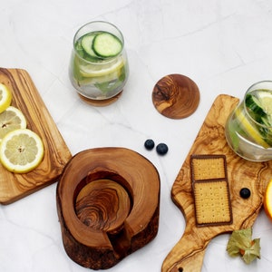 May include: A set of four olive wood serving boards, including a round coaster, a rectangular board with a handle, and two rectangular boards. The boards are arranged on a white surface with lemons, limes, cucumbers, and a glass of water with lemon and cucumber slices.