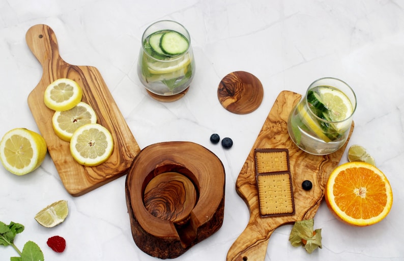 May include: A set of four olive wood serving boards, including a round coaster, a rectangular board with a handle, and two rectangular boards. The boards are arranged on a white surface with lemons, limes, cucumbers, and a glass of water with lemon and cucumber slices.
