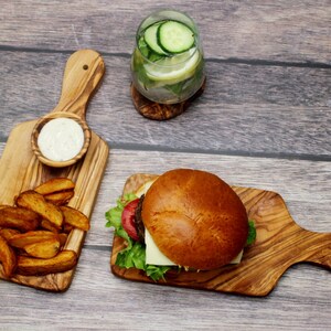 May include: A wooden serving board with a handle, featuring a hamburger with lettuce, tomato, and cheese, and a side of potato wedges. A small bowl of dip is also on the board.