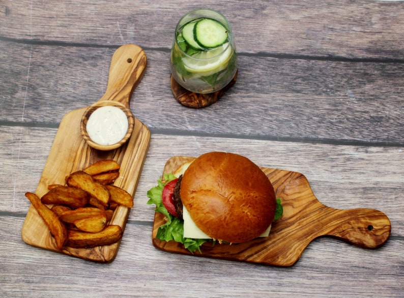 May include: A wooden serving board with a handle, featuring a hamburger with lettuce, tomato, and cheese, and a side of potato wedges. A small bowl of dip is also on the board.
