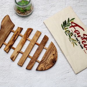 Può includere: Un sottopentola di legno a forma di pesce con un design a lamelle. Il sottopentola è realizzato in legno di ulivo e ha una finitura naturale. Il sottopentola è appoggiato su una superficie bianca.