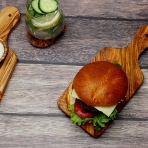 May include: A cheeseburger with lettuce and tomato on a wooden cutting board. There is a side of potato wedges and a bowl of dip on a separate wooden cutting board. A glass of cucumber and lemon water is in the background.