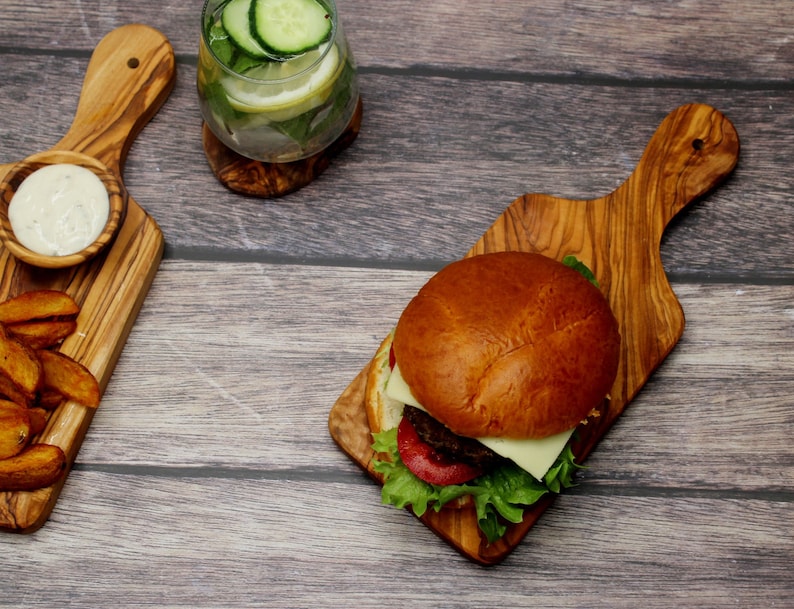May include: A cheeseburger with lettuce and tomato on a wooden cutting board. There is a side of potato wedges and a bowl of dip on a separate wooden cutting board. A glass of cucumber and lemon water is in the background.