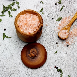 Puede incluir: Un cuenco de madera lleno de sal rosa del Himalaya, con una tapa a juego y una cuchara de madera. La sal es gruesa y tiene un tono rosa claro. Hay granos de pimienta y hierbas secas esparcidas.