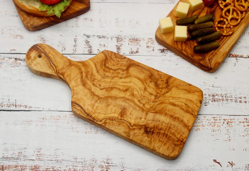 May include: A large, rectangular olive wood cutting board with a handle. The board has a natural wood grain pattern and is perfect for serving cheese, crackers, and other appetizers.