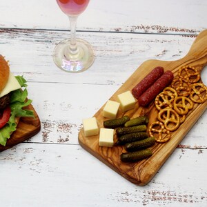 May include: A wooden cutting board with a handle, featuring a variety of snacks, including pretzels, cheese cubes, pickles, and salami.