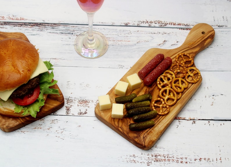 May include: A wooden cutting board with a handle, featuring a variety of snacks, including pretzels, cheese cubes, pickles, and salami.