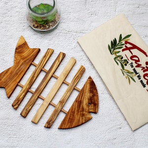 Può includere: Un sottopentola di legno a forma di pesce con una finitura marrone chiaro. Il sottopentola ha un motivo a griglia di listelli di legno. Il sottopentola è su una superficie bianca. Il testo "A-Tech You need We made" è su una borsa di tela bianca.