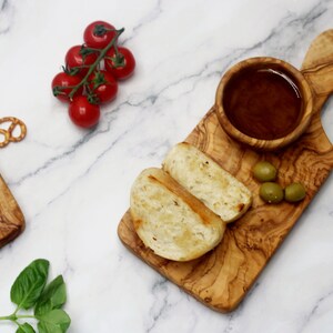 May include: A small olive wood serving board with a handle, a small bowl, two slices of toasted bread, and green olives. The board is on a white marble surface.