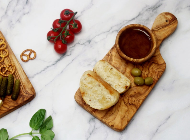 May include: A small olive wood serving board with a handle, a small bowl, two slices of toasted bread, and green olives. The board is on a white marble surface.