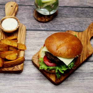 May include: A wooden cutting board with a handle, a burger with lettuce, tomato, and cheese, and a small bowl of dipping sauce. The cutting board is made of olive wood and has a natural grain pattern.
