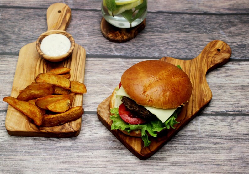 May include: A wooden cutting board with a handle, a burger with lettuce, tomato, and cheese, and a small bowl of dipping sauce. The cutting board is made of olive wood and has a natural grain pattern.