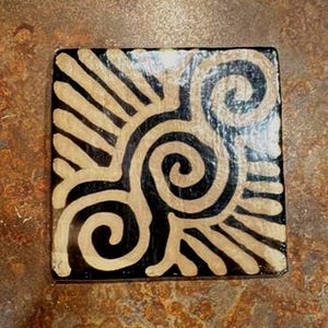 Puede incluir: Posavasos cuadrado con fondo negro y diseño beige estilizado de espiral y rayos de sol. El posavasos tiene un acabado brillante y probablemente está hecho de cerámica o material similar. El diseño es simétrico y abstracto.