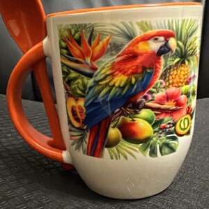 May include: White ceramic mug with an orange handle and spoon. The mug features a colorful parrot illustration with tropical fruits and flowers. The rim and handle are bright orange.