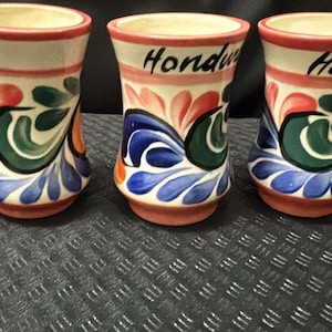 May include: Three ceramic shot glasses with a cream-colored base, featuring colorful floral designs in red, blue, and green. The word "Honduras" is written on two of the glasses, and "A." on the other.