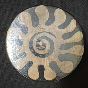 May include: A round, decorative object with a spiral and stylized shapes in a beige and dark gray color scheme. The design is centered on the object, which is wrapped in clear plastic.