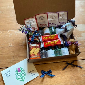 May include: A brown cardboard gift box filled with various items, including tea bags, shortbread biscuits, bath products, and a small stuffed animal. The box is open and the items are visible. The box is decorated with a tartan ribbon and a card that says "Lang May Yer Lum Reek!" and "With Love from Scotland".