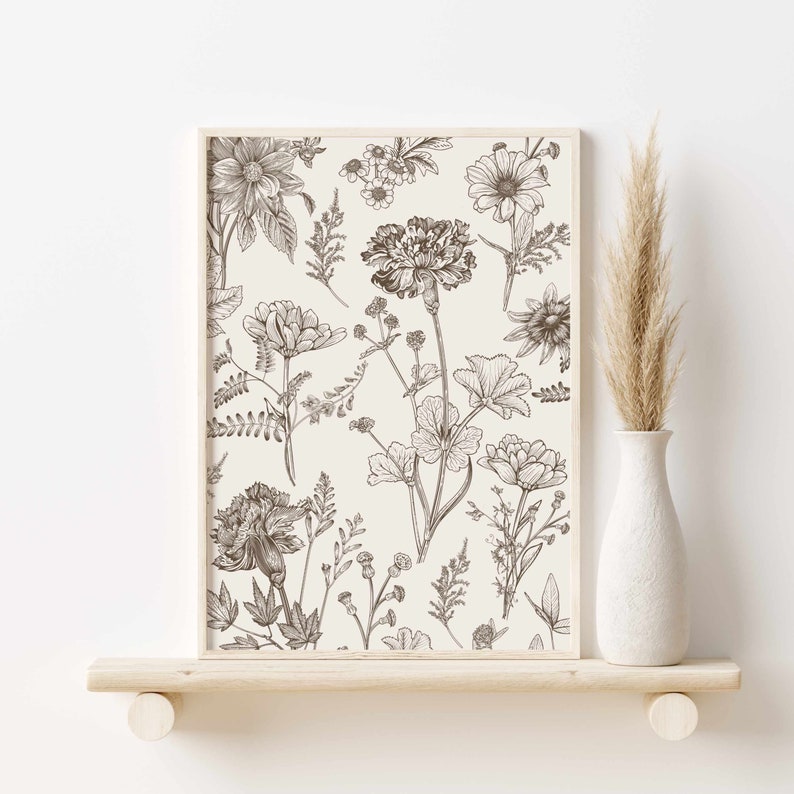 May include: A framed botanical print featuring detailed line drawings of various flowers and foliage in shades of brown and beige. The artwork is displayed on a light wooden shelf, with a white vase holding dried pampas grass to the right.