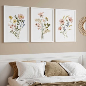 May include: Three framed floral art prints with watercolor flower arrangements in white frames. The flowers are in shades of pink, yellow, and blue. A round woven mirror hangs on the wall. A bed with white pillows and a brown blanket is in the foreground.
