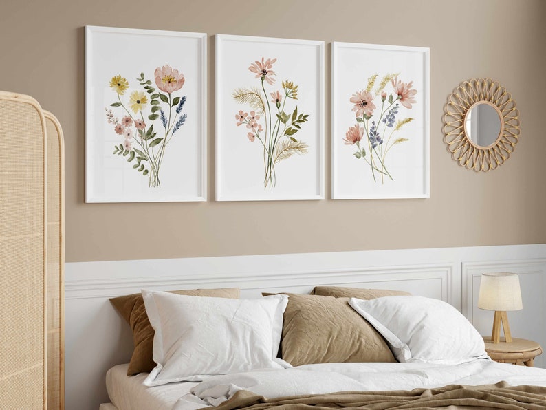 May include: Three framed floral art prints with watercolor flower arrangements in white frames. The flowers are in shades of pink, yellow, and blue. A round woven mirror hangs on the wall. A bed with white pillows and a brown blanket is in the foreground.