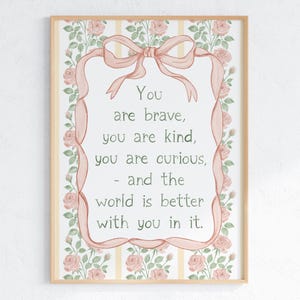 May include: A framed print with a light wood frame. The artwork features a pink ribbon and floral border, with the words "You are brave, you are kind, you are curious, - and the world is better with you in it." in the center.
