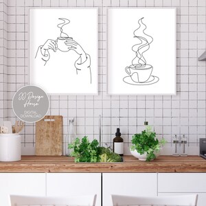 May include: Two black and white line art prints of a coffee cup with steam rising from it. The first print shows a hand holding the cup, while the second print shows the cup on a saucer. The prints are framed in white and hung on a white tiled wall.