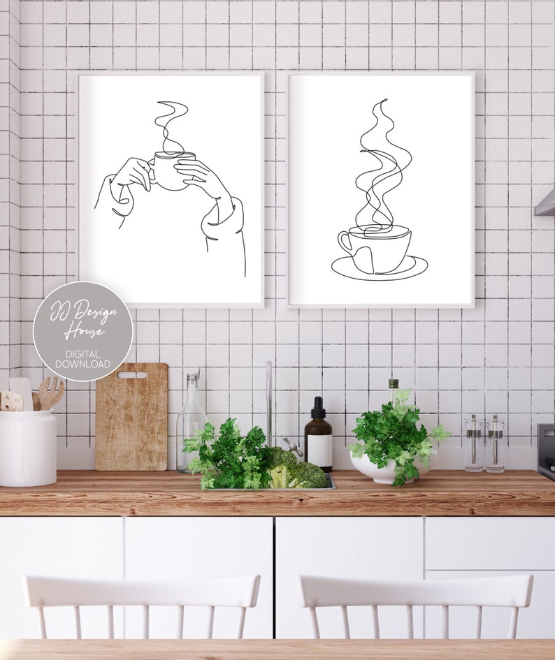 May include: Two black and white line art prints of a coffee cup with steam rising from it. The first print shows a hand holding the cup, while the second print shows the cup on a saucer. The prints are framed in white and hung on a white tiled wall.