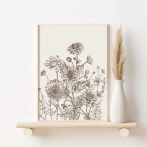 May include: A framed botanical print featuring a detailed pen and ink illustration of various flowers and foliage. The artwork is in shades of brown and cream, with a minimalist aesthetic. A white vase with dried pampas grass sits on a wooden shelf.
