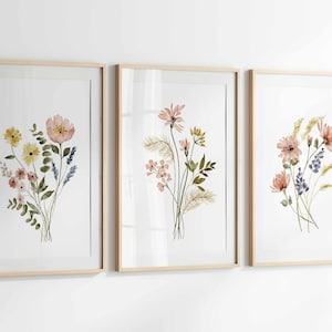 May include: Three framed watercolor prints featuring floral arrangements. Each print showcases a different bouquet of flowers in shades of pink, yellow, and blue, with green foliage. The frames are a light wood color, and the background is white.