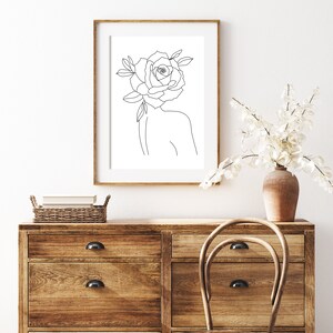 May include: A framed art print featuring a minimalist line drawing of a person with a rose for a head. The artwork is in a wooden frame and is displayed on a wooden dresser. A vase with white flowers and a wicker basket are also visible.